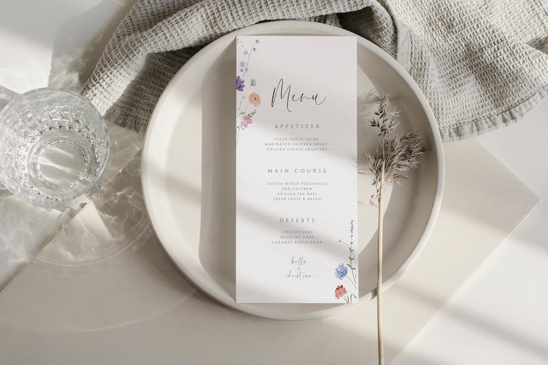 May include: A white menu card with a floral design and the text "Menu", "Appetizer", "Field Green Salad", "Marinated Chicken Breast", "Grilled Cheese Cro&ucirc;tons", "Main Course", "Roasted Mixed Vegetables", "Grilled Sea Bass", "Fresh Fruit & Bread", "Desserts", "Cheesecake", "Wedding Cake", "Caramel Apple Crisp", "Belle + Christian" printed on it.