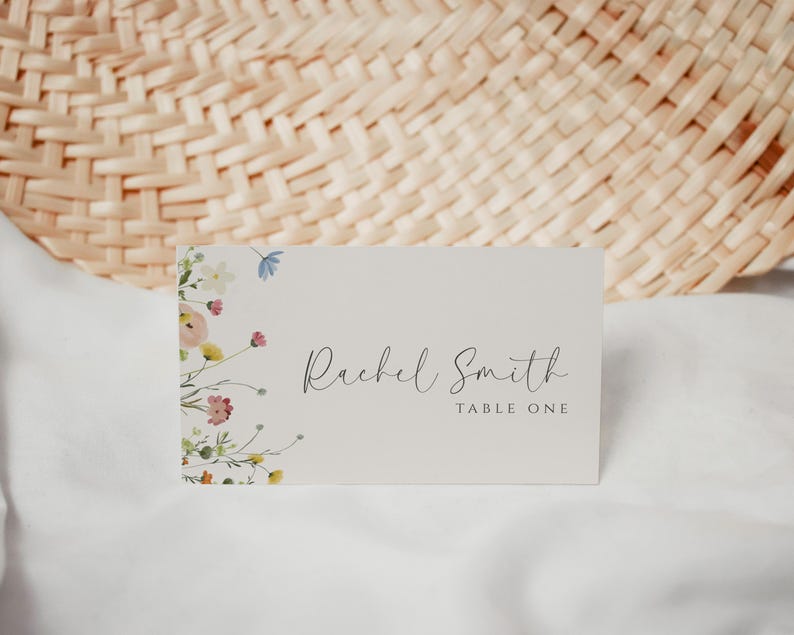 May include: A white table card with a floral design on the left side and the text "Rachel Smith Table One" on the right side.