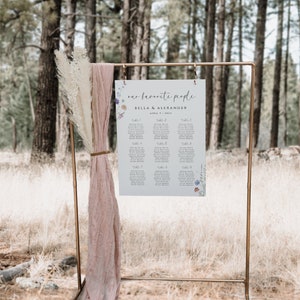 May include: A wedding seating chart with a floral design. The chart is printed on white paper and has a light blue background. The chart is titled "our favorite people" and lists the names of guests and their assigned tables. The chart is displayed on a gold metal stand in a forest setting.