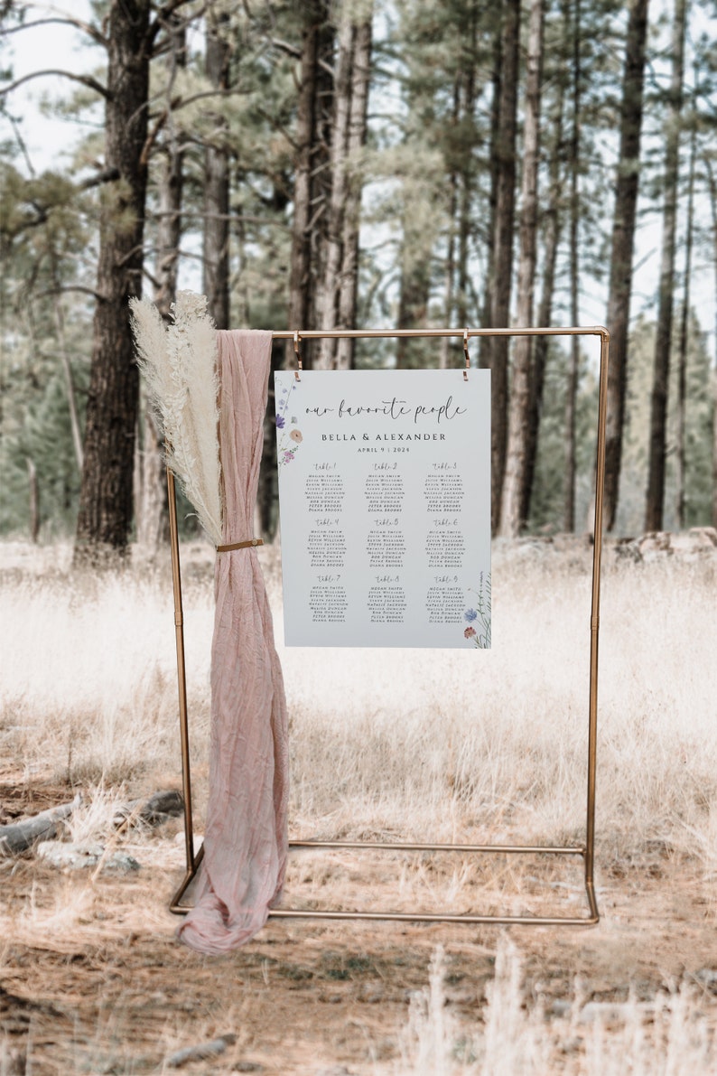 May include: A wedding seating chart with a floral design. The chart is printed on white paper and has a light blue background. The chart is titled "our favorite people" and lists the names of guests and their assigned tables. The chart is displayed on a gold metal stand in a forest setting.