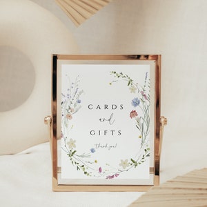 May include: A gold-framed sign with a white background and a floral wreath design. The sign reads "Cards and Gifts, thank you!"