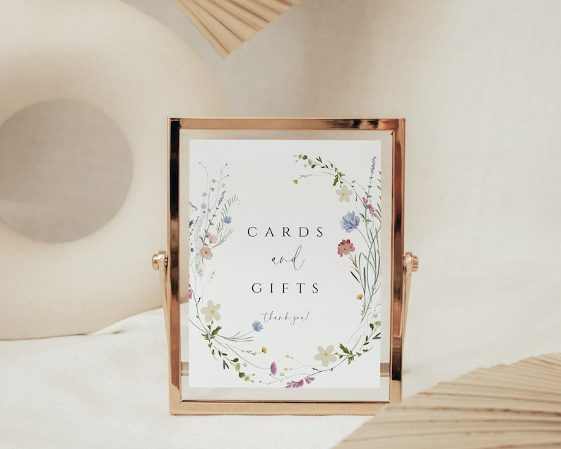 May include: A gold-framed sign with a white background and a floral wreath design. The sign reads "Cards and Gifts, thank you!"
