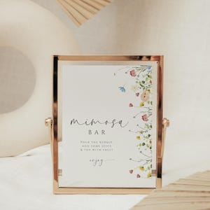 May include: A gold-framed sign with a floral border and the text "mimosa bar" in cursive. The sign also includes instructions for making a mimosa: "Pour the bubbly, add some juice & top with fruit, enjoy."