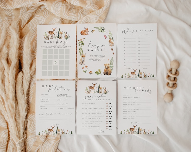 May include: A collection of baby shower game cards with woodland animal illustrations. The cards include "Baby Bingo", "Diaper Raffle", "Who's That Baby?", "Baby Predictions", "Guess Who Mommy or Daddy?", and "Wishes for Baby". A wooden rattle is also present.