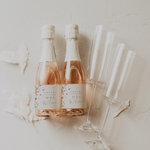 May include: Two mini bottles of champagne with floral labels that say "Cheers to the future Mrs. Wilson". Two champagne flutes are also in the image.