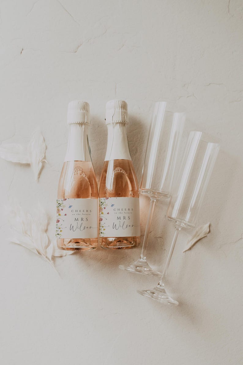 May include: Two mini bottles of champagne with floral labels that say "Cheers to the future Mrs. Wilson". Two champagne flutes are also in the image.