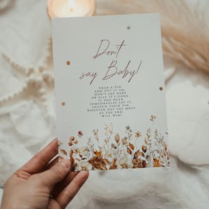May include: A rectangular card with the text "Don't say Baby!" in a handwritten font. The card has a floral design at the bottom and is held by a hand. The card's instructions are: Grab a pin and put it on. Don't say baby or else it's gone.
