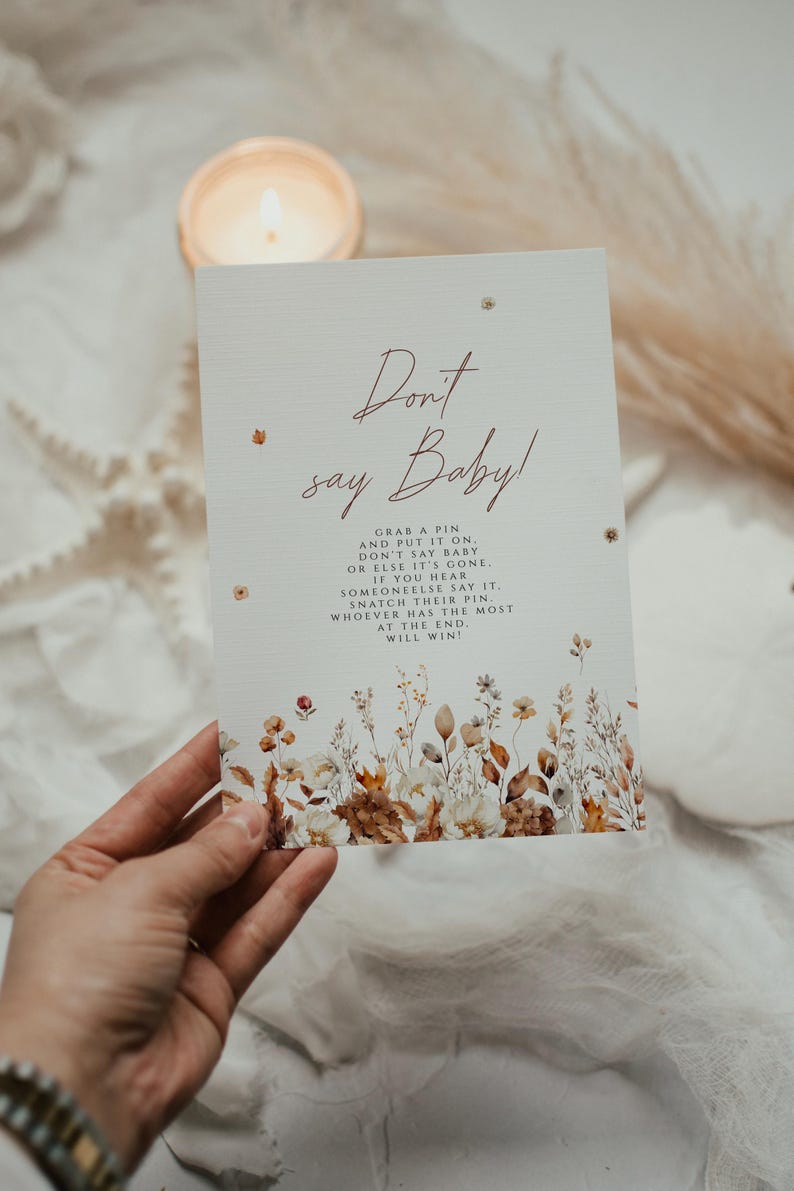 May include: A rectangular card with the text "Don't say Baby!" in a handwritten font. The card has a floral design at the bottom and is held by a hand. The card's instructions are: Grab a pin and put it on. Don't say baby or else it's gone.