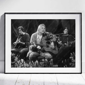 May include: Monochrome framed print depicting three musicians playing acoustic guitars. The image shows the musicians seated, with one in the center singing. The print is displayed on a white wall above a wooden floor.