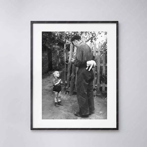 May include: A black and white photograph of an older man holding a small white piglet in his arms. He is standing next to a young boy who is looking up at him. The man is wearing a striped shirt and a cap. The boy is wearing a short-sleeved shirt and shorts. They are standing in front of a wooden fence.