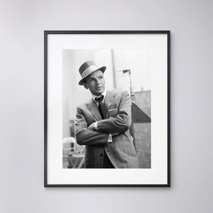 Pu&ograve; includere: Fotografia in bianco e nero di un uomo in abito e cappello. &Egrave; in piedi con le braccia incrociate e guarda di lato.