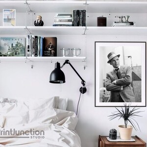 Pu&ograve; includere: Fotografia in bianco e nero di un uomo con un cappello fedora e un abito. L'uomo guarda di lato e ha un'espressione seria sul viso. La fotografia &egrave; incorniciata in una cornice nera e appesa a un muro bianco.