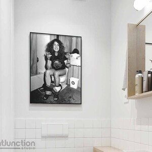 May include: Black and white photo of a person sitting on a toilet, smoking a cigarette. The photo is framed and hanging on a white tiled wall.
