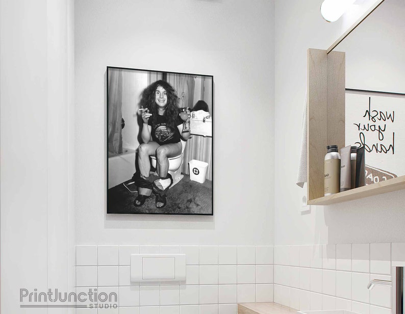 May include: Black and white photo of a person sitting on a toilet, smoking a cigarette. The photo is framed and hanging on a white tiled wall.