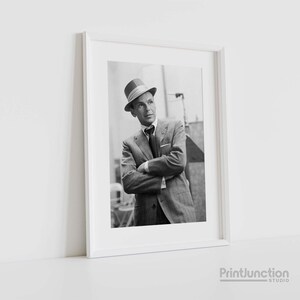 Pu&ograve; includere: Fotografia in bianco e nero di un uomo in abito e cappello. &Egrave; in piedi con le braccia incrociate e guarda in alto.