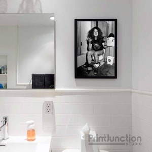 May include: Black and white photo of a person sitting on a toilet. The photo is framed in a black frame and is hanging on a white tiled wall.