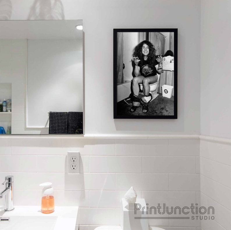 May include: Black and white photo of a person sitting on a toilet. The photo is framed in a black frame and is hanging on a white tiled wall.