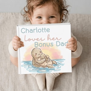 Może przedstawiać: Niemowlę trzyma książkę z napisem "Charlotte Loves her Bonus Dad". Książka zawiera ilustrację dw&oacute;ch kresk&oacute;wkowych ż&oacute;łwi w wodzie z tłem zachodu słońca. Okładka książki ma białą obw&oacute;dkę.
