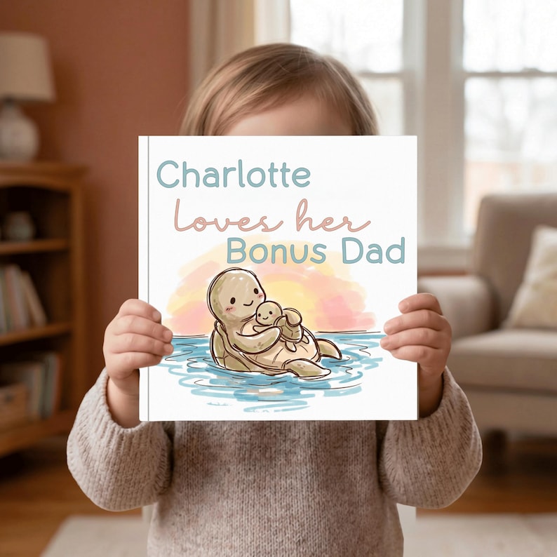Może przedstawiać: Książka dla dzieci z tekstem "Charlotte Loves her Bonus Dad" w niebieskich i r&oacute;żowych literach. Okładka przedstawia ilustrację ż&oacute;łwia obejmującego ż&oacute;łwia niemowlęcego w wodzie z tłem zachodu słońca. Książka jest trzymana przez osobę.