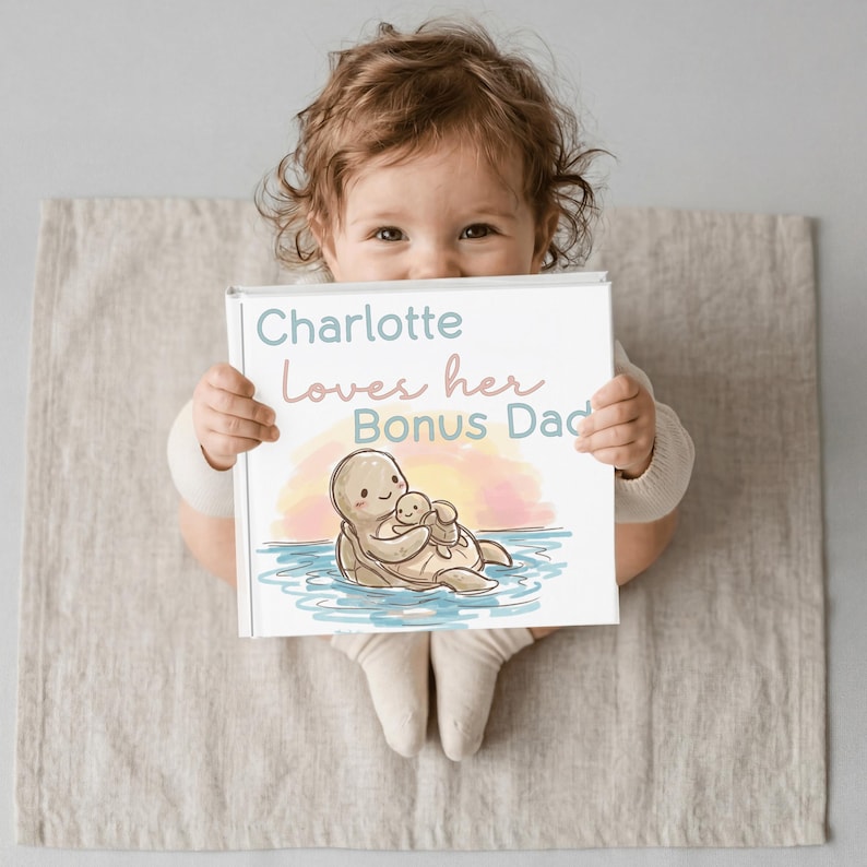 Może przedstawiać: Niemowlę trzyma książkę z napisem "Charlotte Loves her Bonus Dad". Książka zawiera ilustrację dw&oacute;ch kresk&oacute;wkowych ż&oacute;łwi w wodzie z tłem zachodu słońca. Okładka książki ma białą obw&oacute;dkę.