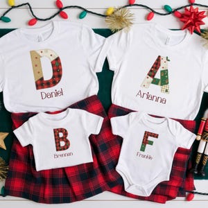 May include: White t-shirts and a babygrow with initial letters in a patchwork design. The letters feature red and green plaid patterns, with names printed below. The shirts are paired with red and black plaid skirts.