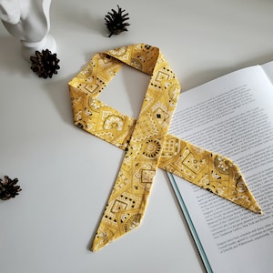Könnte beinhalten: Ein gelbes Bandana mit einem schwarzen und weißen Paisley-Muster. Das Bandana ist zu einem Knoten gebunden und liegt auf einem Buch.