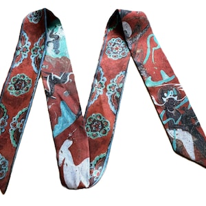 May include: A long, narrow scarf in a deep rust-red colour featuring a repeating pattern of stylised floral motifs in teal and white, alongside graphic illustrations of white deer and dark figures set against the red background.