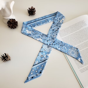May include: A light blue bandana with a white and black paisley pattern. The bandana is tied in a bow and is lying on a white surface.