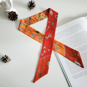 Puede incluir: Una bufanda de seda roja y naranja con un patrón de llaves y borlas. La bufanda está doblada en un bucle y está sobre un libro.
