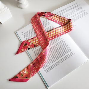 May include: A pink and yellow patterned silk scarf with a geometric design. The scarf is tied in a knot and is lying on a white book.