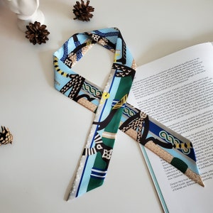 May include: A blue and green patterned silk scarf with a geometric design. The scarf is folded and tied in a knot, and the ends are hanging down. The scarf is on a white surface with a book underneath it.