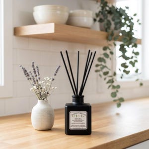 May include: A black square essential oil diffuser with black reeds and a label that reads "MADE PURE CO MINTA". A small white vase with dried lavender and white flowers sits to the left. The items are on a wooden countertop.
