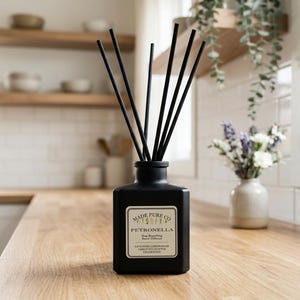 May include: A black square essential oil diffuser with black reeds. The label reads "MADE PURE CO. PETRONELLA Bug Repelling Reed Diffuser". The diffuser sits on a light wood surface. A small vase of flowers is in the background.