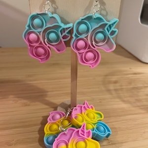 Könnte beinhalten: Ein Paar Ohrringe in Form eines Einhorns mit einem Pop-it-Design. Die Ohrringe sind aus Silikon und haben ein Farbkonzept in Pink, Blau und Gelb.