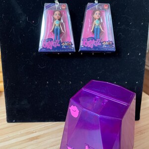 以下が含まれることがあります： Bratz人形のイヤリング。それぞれクリアケースに入ったミニチュアの人形が特徴です。イヤリングはシルバーカラーです。紫色の幾何学的なMini Bratzコンテナも存在します。