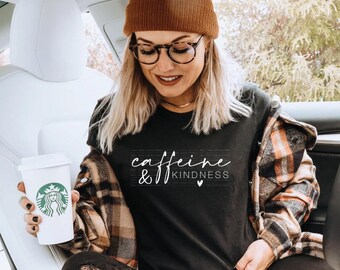May include: A black t-shirt featuring the white text "caffeine & KINDNESS" with a small heart icon. The shirt is worn with a brown knit beanie, a brown and black plaid flannel shirt, and black distressed jeans, while holding a white coffee cup.
