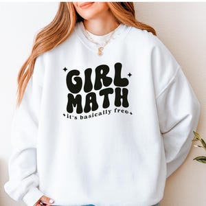 Puede incluir: Una sudadera blanca de cuello redondo con el texto en negro GIRL MATH y la frase it's basically free en una tipograf&iacute;a ondulada de estilo retro. El dise&ntilde;o est&aacute; decorado con peque&ntilde;as estrellas de cuatro puntas alrededor de las letras.