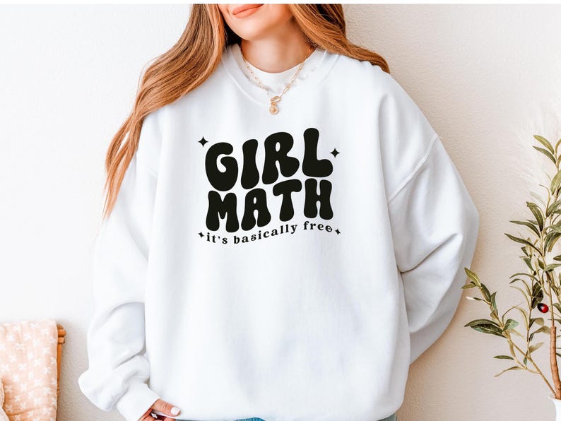 Puede incluir: Una sudadera blanca de cuello redondo con el texto en negro GIRL MATH y la frase it's basically free en una tipograf&iacute;a ondulada de estilo retro. El dise&ntilde;o est&aacute; decorado con peque&ntilde;as estrellas de cuatro puntas alrededor de las letras.