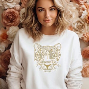 May include: A white sweatshirt with a gold leopard design. The design includes the word "TIRED" and the phrase "POUR SOME COFFEE ON ME". The sweatshirt is a crew neck style.