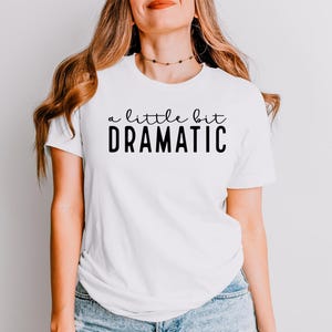 May include: A white short-sleeved t-shirt featuring the black text a little bit DRAMATIC printed on the chest. The shirt has a crew neckline and is paired with blue denim jeans. The design uses a mix of cursive and bold sans-serif fonts.