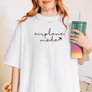 Peut inclure: T-shirt blanc avec l'inscription "airplane mode" en &eacute;criture noire et un petit avion. Un grand gobelet transparent avec une paille est tenu dans la main. Le gobelet pr&eacute;sente un d&eacute;grad&eacute; de couleurs, du jaune au rose et au bleu.