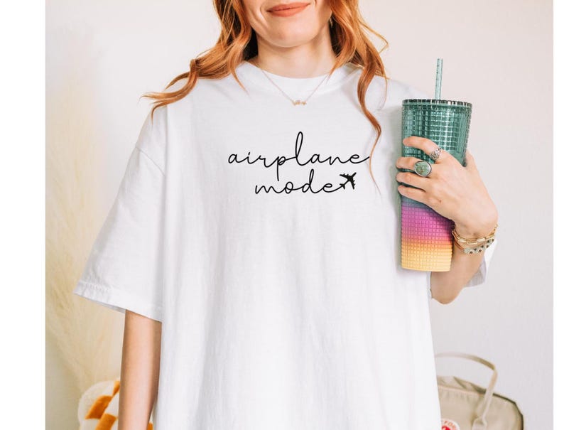 Peut inclure: T-shirt blanc avec l'inscription "airplane mode" en &eacute;criture noire et un petit avion. Un grand gobelet transparent avec une paille est tenu dans la main. Le gobelet pr&eacute;sente un d&eacute;grad&eacute; de couleurs, du jaune au rose et au bleu.