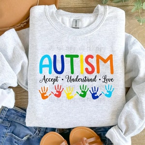 May include: A gray sweatshirt with the word "AUTISM" in rainbow colors and the text "Accept. Understand. Love." below it. There are six rainbow handprints below the text.