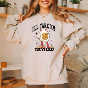 May include: Cream-colored sweatshirt with the text "I'll Take 'Em" above a graphic of a deviled egg character with horns, a tail, and boots. The word "DEVILED" is below the graphic, with flames at the bottom.