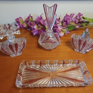 Op de afbeelding: Een vintage toilettafelset met heldere glazen parfumflesjes en een bijpassend dienblad. De set heeft rode accenten en ingewikkelde geslepen glasontwerpen. De flesjes hebben unieke stoppers en het dienblad heeft een zonnepatroon.