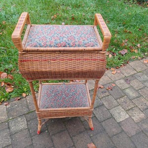 以下が含まれることがあります： 上部に花柄のクッション、下部に小さな籐製の棚が付いた籐製の収納オットマン。オットマンは赤色のアクセントがついた茶色で、レンガの敷き詰められたパティオの上に置かれています。