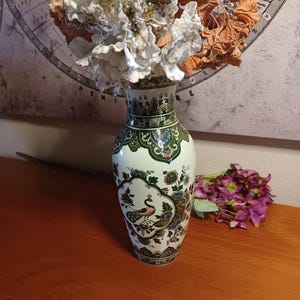 Op de afbeelding: Een witte keramische vaas met een pauwenontwerp, groene en gouden accenten en een bloemstuk. De vaas is ongeveer 30 cm hoog. De achtergrond bevat een wereldkaart en een boeket paarse bloemen.