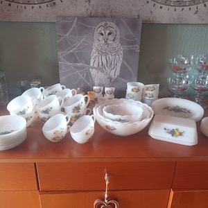 May include: A collection of white milk glass dinnerware featuring a fruit and grape pattern, including multiple teacups, saucers, nested scalloped bowls, a rectangular butter dish, and a set of clear glass goblets with red accents on the stems.