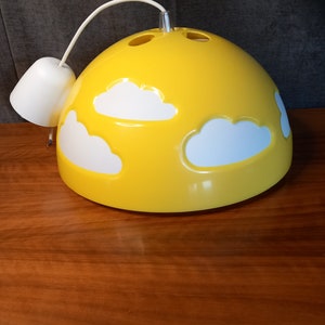 May include: A yellow pendant light with white cloud cutouts. The light fixture has a rounded, dome-like shape and a white cord and ceiling mount. The lamp is sitting on a wooden surface.