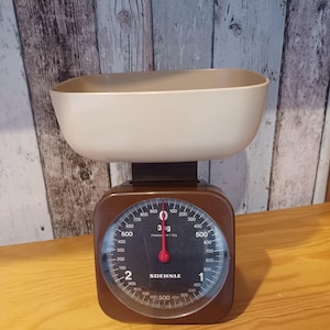 Op de afbeelding: Een vintage bruine keukenweegschaal met een crèmekleurige kom. De wijzerplaat van de weegschaal heeft een ronde wijzerplaat met zwarte cijfers en een rode naald. De merknaam "SOEHNLE" staat op de wijzerplaat.